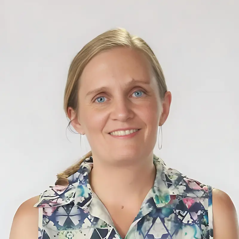 Freya with blonde hair pulled back smiles at the camera. She is wearing a sleeveless top with a geometric pattern and hoop earrings. The background is plain and light-colored.