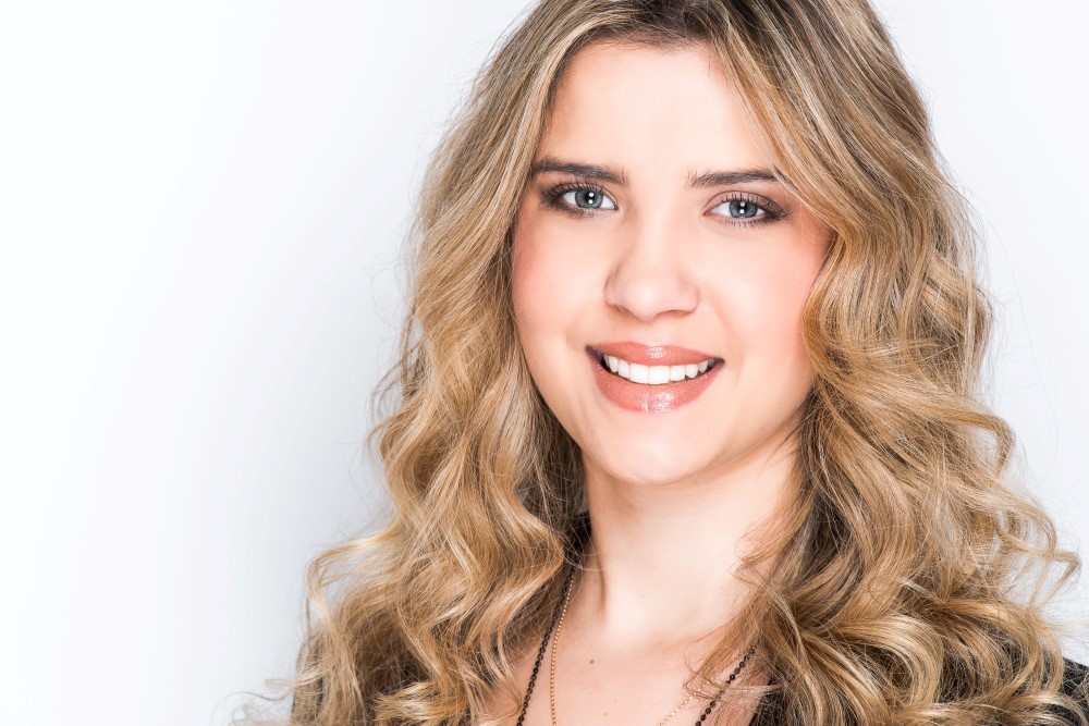 Rachael with long, curly blonde hair smiles at the camera. She is wearing a black top and is posed against a plain white background.