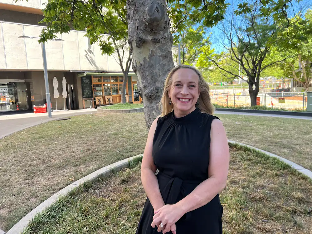 A smiling woman with blonde hair, wearing a black sleeveless dress, stands with her hands clasped next to a tree in a grassy outdoor area, with a building and walkway visible in the background.