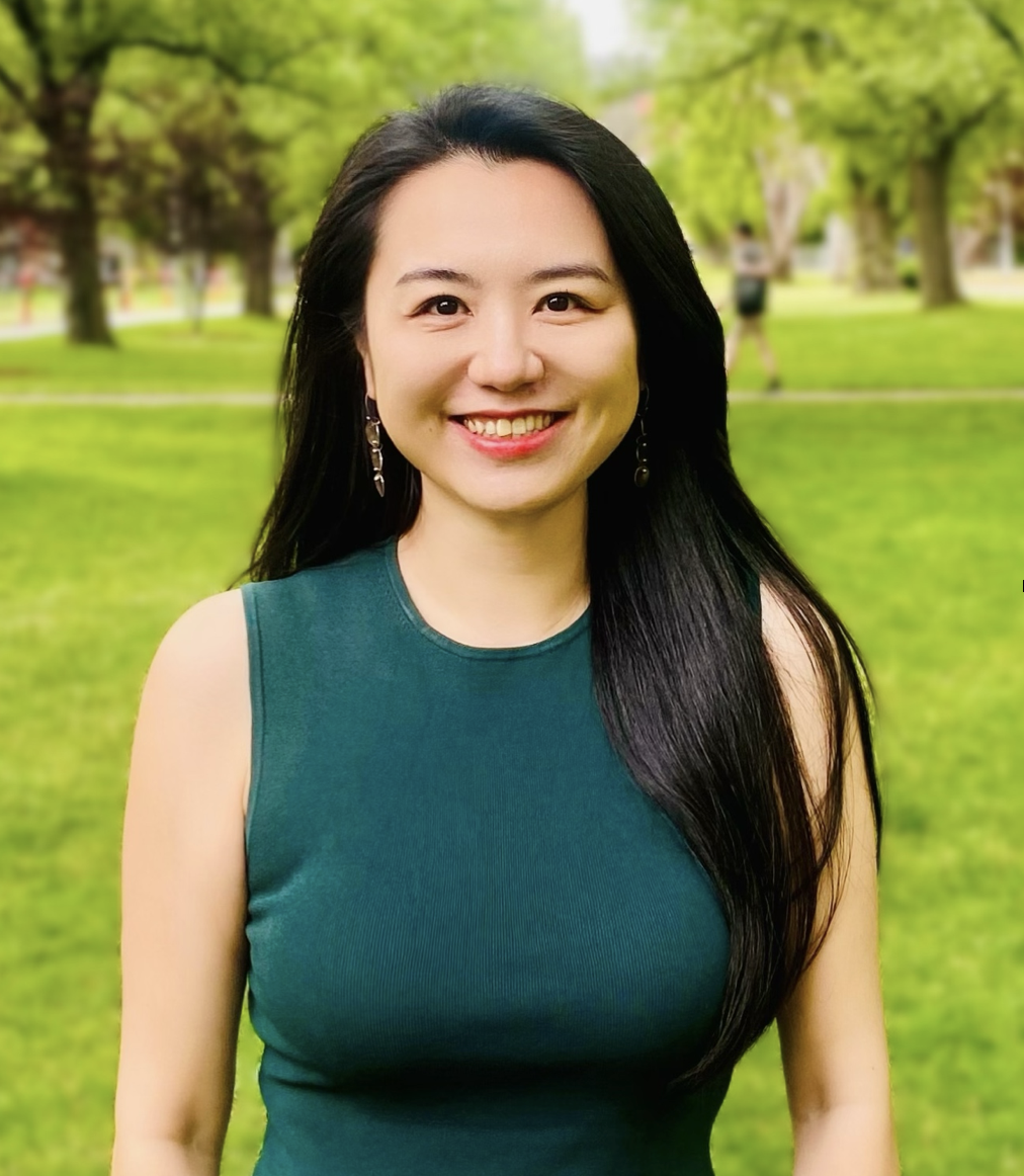 Lyn with long black hair and wearing a sleeveless green top smiles at the camera while standing in a park with green grass and trees in the background.